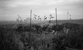 A grayscale photograph of the phrase "The Day-Sob-Dies" suspended between two thin poles in an overgrown field against the skyline of a city in the distance.