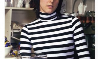 A color photograph of a young woman with light skin and dark hair in a black-and-white striped turtleneck standing behind a cafe counter.