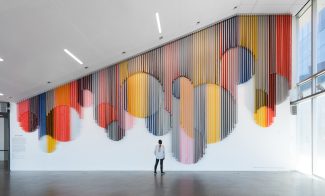 A visitor standing in a spacious lobby and looking up at a monumental hanging wall sculpture made from bands of colorful coated mesh fabric draped in various lengths to create series of interlocking circular forms.