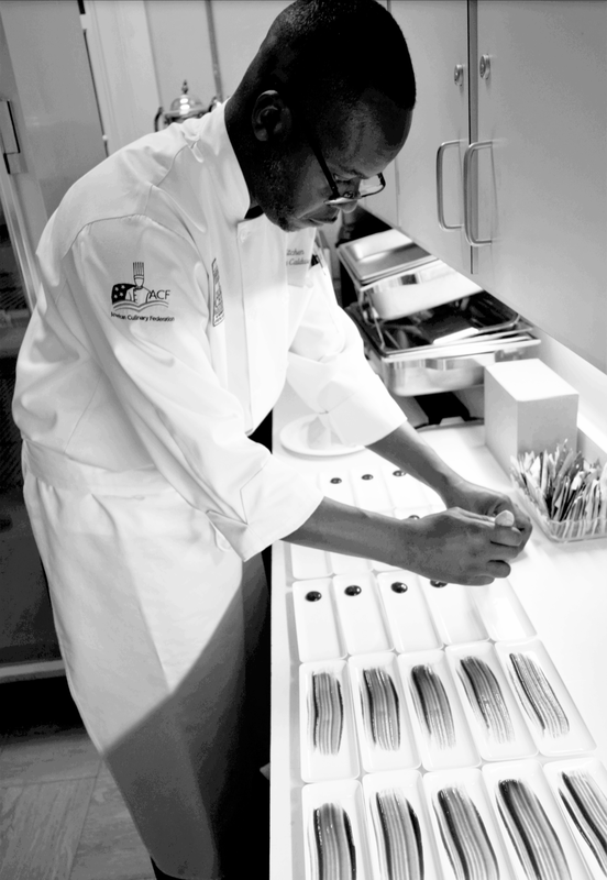 Black and white photo of bald man in a chef's jacket plating many dishes