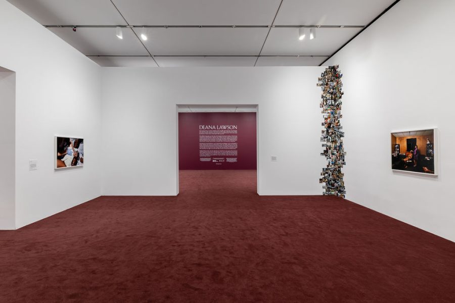 Gallery view of framed photos hanging on the wall with an assemblage of photos in the right corner. The next room is visible through the entry way, showing a purple painted wall with wall text introducing the exhibition.