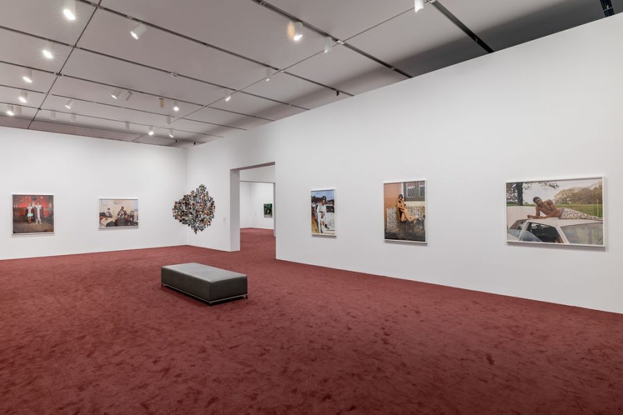 A wide shot of a gallery room with wine colored carpet and several large format photos framed and hung. In the corner is an assemblage installation of photographs.