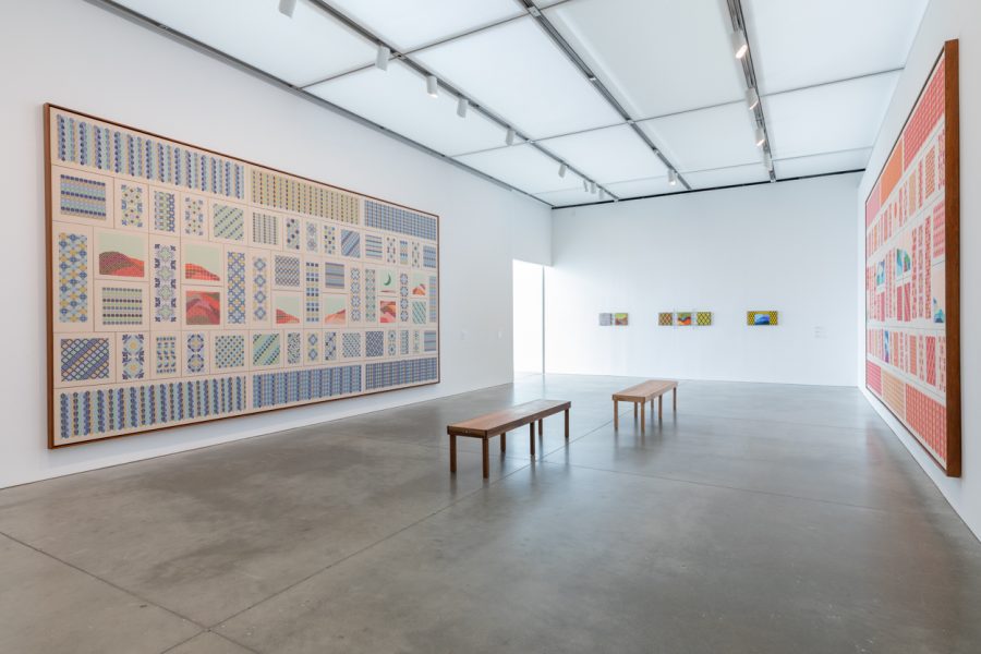 Gallery with two large rectangular embroideries hanging opposite each other with wooden benches in the middle
