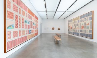 Large, framed embroidered work consisting of patterned rectangles hang opposing each other in a gallery with benches between them