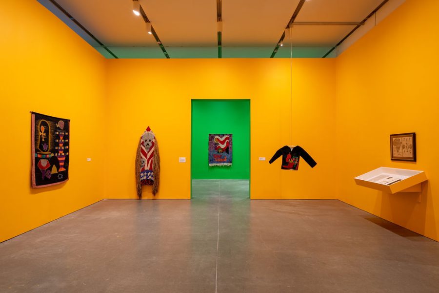 An orange-walled gallery with a few textile works, including a shirt hanging from the ceiling. The next gallery is painted green with a colorful textile work in view.