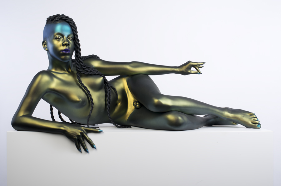 A life-size, hyper realistic bronze sculpture of a nude with long, thick hair in twists, lying on their side, propped up on one elbow with the other hand extended. The finish is a metallic green, except for blue fingernails and purple lipstick.