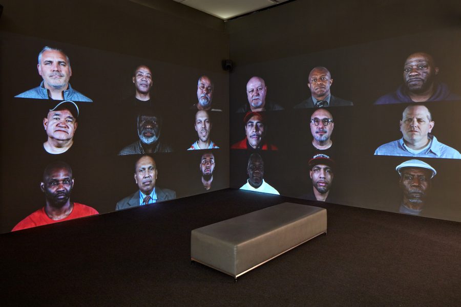 Two adjacent wall-sized screens in a darkened room show a grid of video portraits of men.