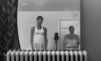 A black-and-white photograph shows the artist, a Black woman, and her mother in lounge wear and hair caps reflected in a mirror resting on a gas radiator.