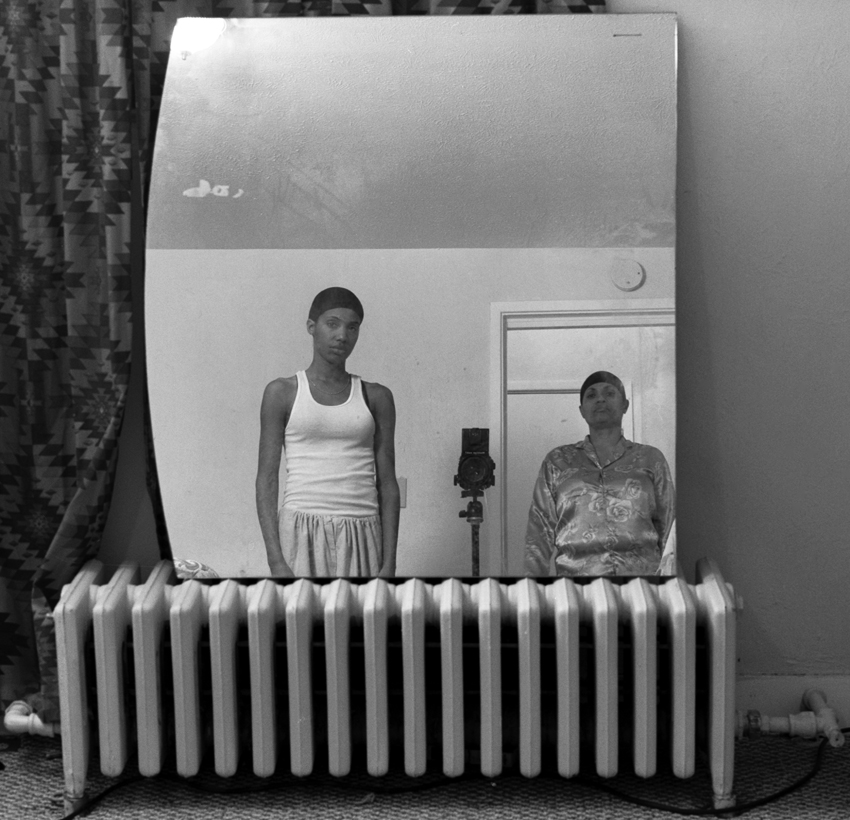 A black-and-white photograph shows the artist, a Black woman, and her mother in lounge wear and hair caps reflected in a mirror resting on a gas radiator.