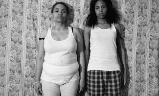A black-and-white photograph shows the artist, a Black woman, standing shoulder to shoulder with her mother before floral-patterned wallpaper. They are wearing white tank tops and casual bottoms and gaze directly at the viewer.