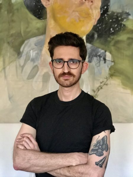 A man in glasses and a black t-shirt in front of a painting. 