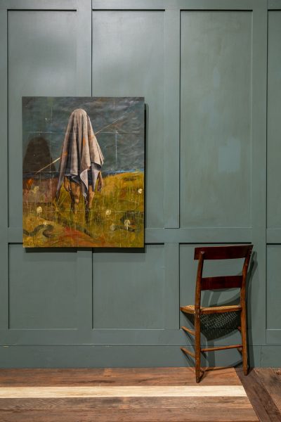 Painting of a figure with a sheet over their head standing in a field of grass hangs on a painted robin's blue paneled wall with a wooden chair installed going through the wall