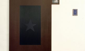 An installation consists of an oil painting of a black flag with a grey star, a much smaller black-and-white photograph of a couple in military uniform, and a blue book jacket with a white star..
