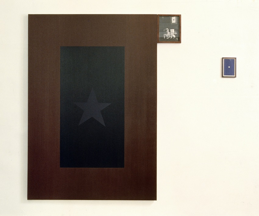 An installation consists of an oil painting of a black flag with a grey star, a much smaller black-and-white photograph of a couple in military uniform, and a blue book jacket with a white star..