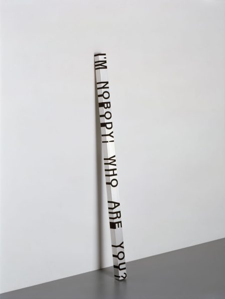 A sculpture depicts a rectangular white pole leaning against a white wall with black text reading, 