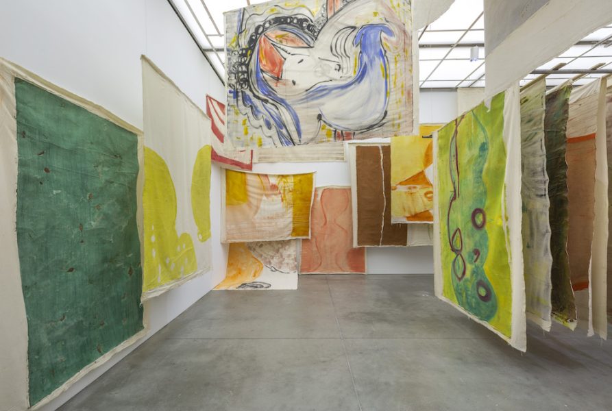 A photograph shows a room full of colorful unframed canvases hung from the ceiling at different angles.