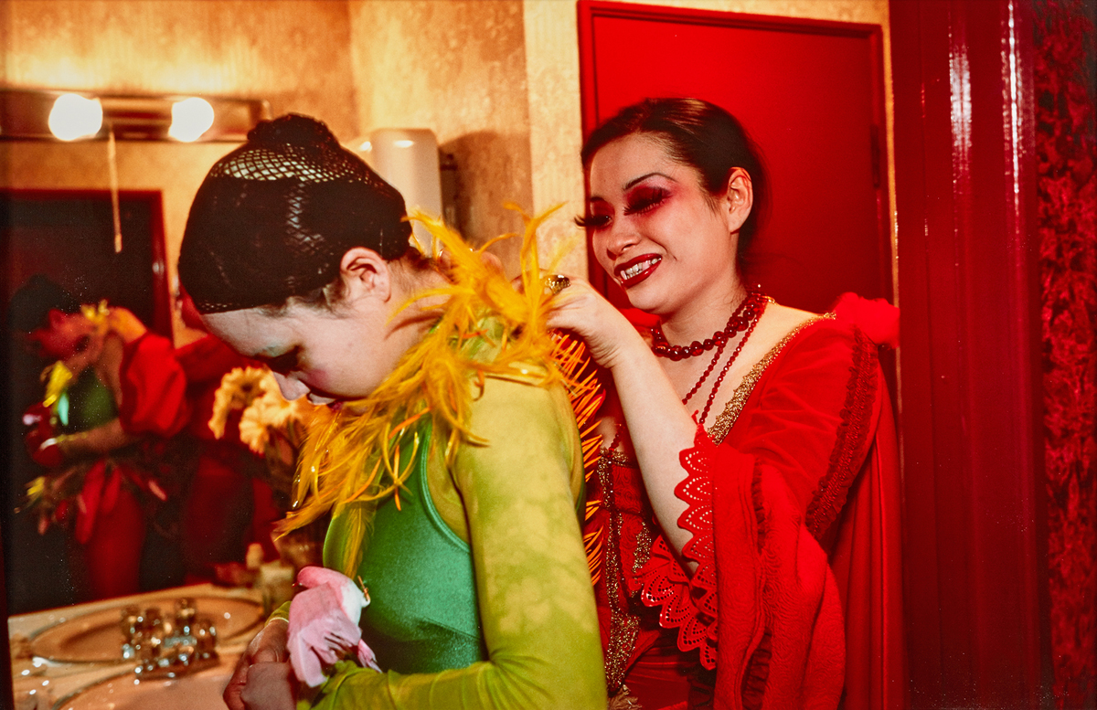 A color photograph of two heavily made-up performers in red and green feathered evening gowns helping each other get ready in a dressing room.