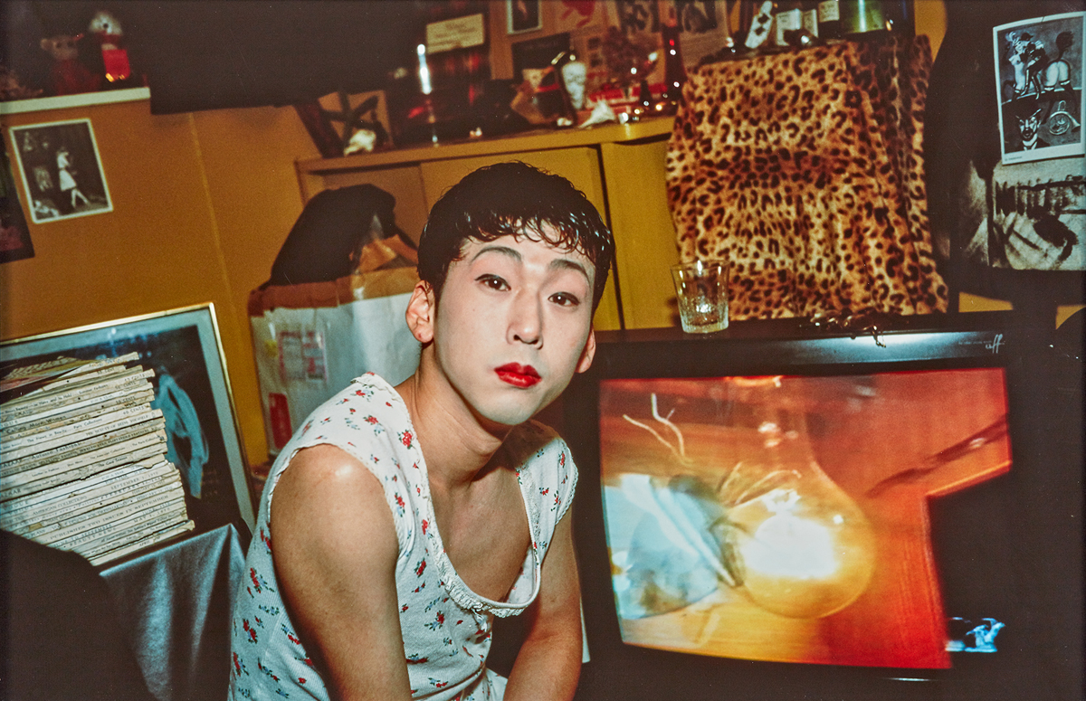 A color photograph of a Japanese adult with powdered makeup and red lipstick looking directly at the viewer in a cluttered room.