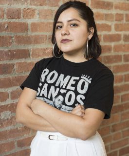A headshot of Xlmena Izquierdo Ugaz wearing a Romeo Santos t-shirt in front of a brick wall.