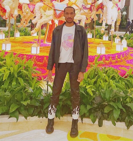 A stylishly-dressed African-American man standing in front of a colorful, textured carousel with horses.