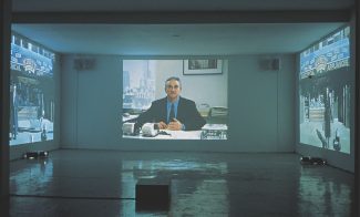 An installation of three videos projected onto three adjacent walls of a gallery. The central image shows a middle-aged white man in a suit seated in an office looking directly at the viewer, the others show a city storefront.