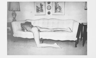 A grainy black-and-white photograph of a pale nude figure lounging face-down on a couch, their left leg hanging down and resting on the floor.
