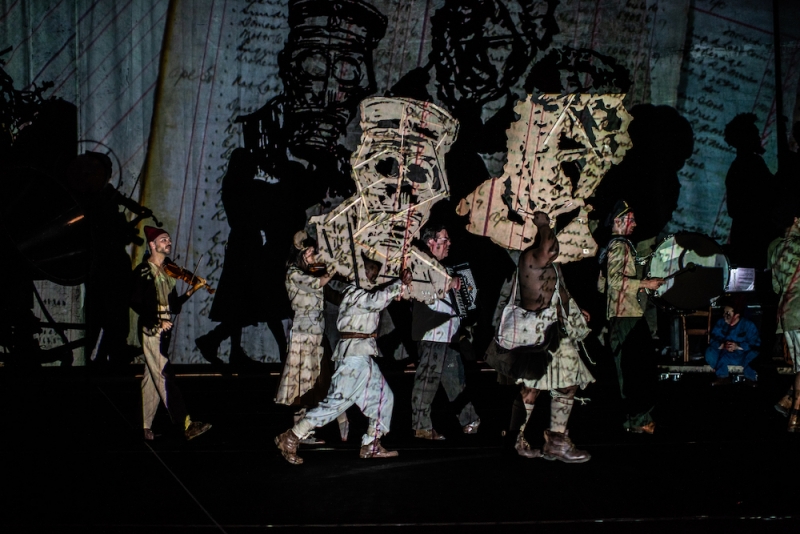 Several people on a stage carry larger-than-life cutouts of drawn heads. An image is projected onto them. 