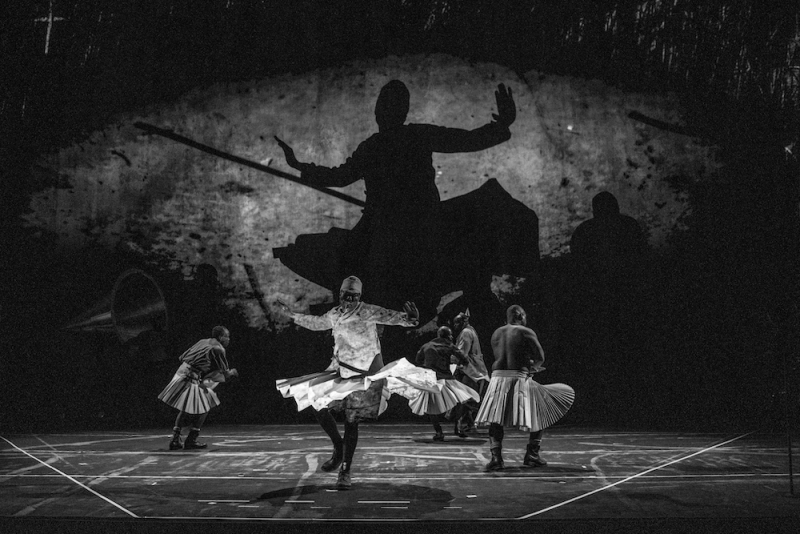 A black and white image of several dancers in pleated white skirts, not in unison. A backdrop shows the enlarged silhouette of one.