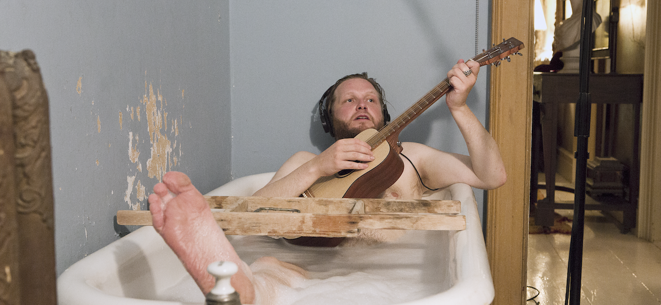 A person wearing headphones plays a guitar while sitting in a bathtub filled with bubbles. A wooden plank rests across the tub. The room has a rustic appearance with peeling paint on the walls. A microphone stand is visible nearby.