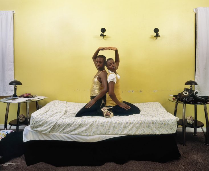 In this photo two adolescent girls kneel back to back on an unmade bed, looking at the camera. They are wearing black pants and gold tops and each have one arm stretched up and touching the other's hand. The wall behind them is yellow with scuff marks. The sheets are white with yellow flowers.