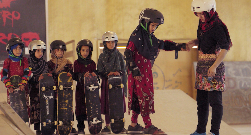 A group of girls wearing helmets and headscarves standing in a line holding skateboards.