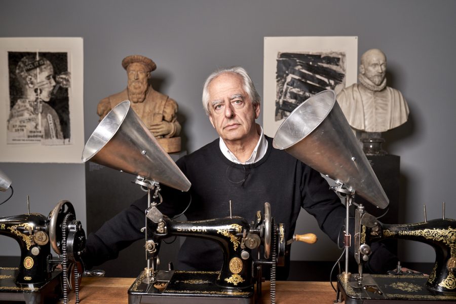A portrait of artist William Kentridge, an elderly white man, standing behind a table with antique sewing machines and silver megaphones attached to them; behind him are classical bust sculptures and prints.