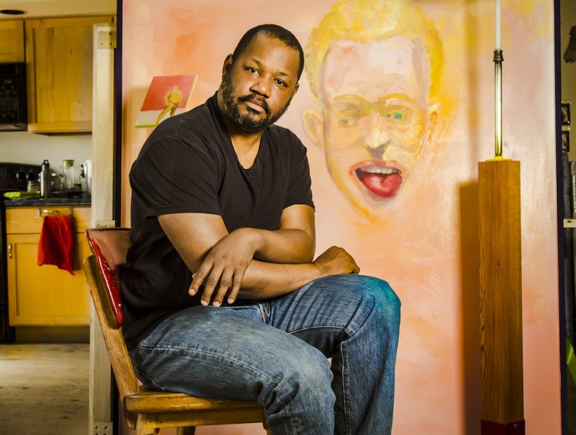 Artist Steve Locke sits in his studio in front of a large painting of a face with its tongue sticking out