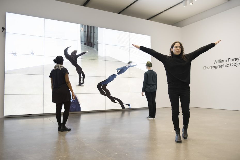 Three women in dark clothing move around a room lined on one side by mirrors that distort their reflections. In the foreground, a young woman faces the viewer with arms stretched in a Y position.