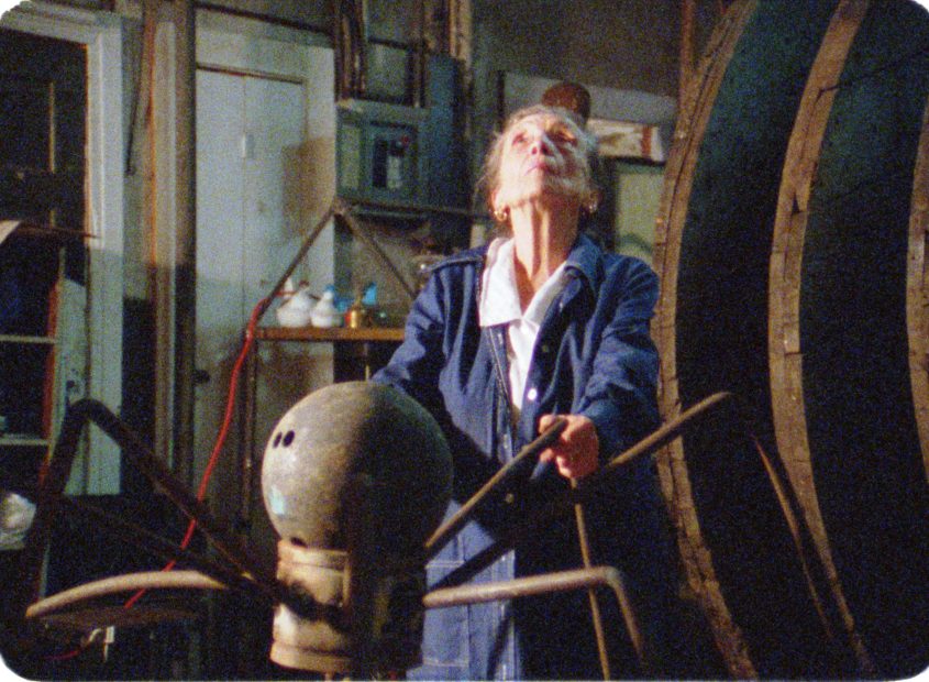 Inside an industrial studio, an elderly white woman looks up as she holds onto spider legs-like pipes from a metal sculpture.