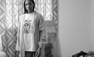 A black-and-white photograph shows the artist, a Black woman, gazing directly at the viewer with the reflection of her mother visible in a mirror behind her. She is wearing a T-shirt with the cast of the Cosby Show on it.