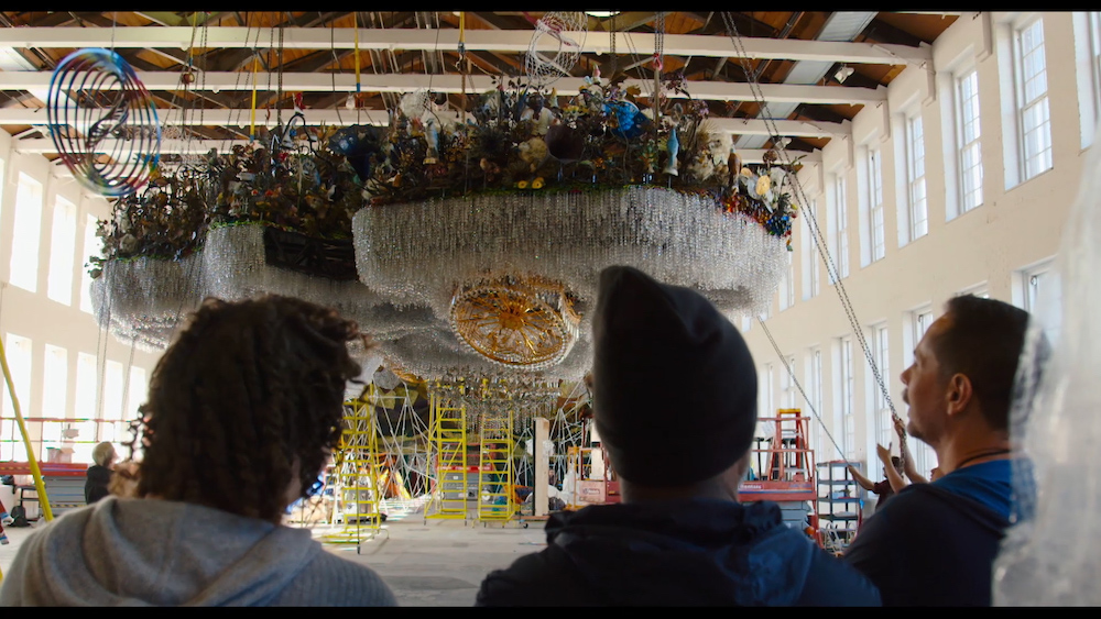Art handlers appear to hoist a massive installation made of beads and sundry objects up toward the ceiling.