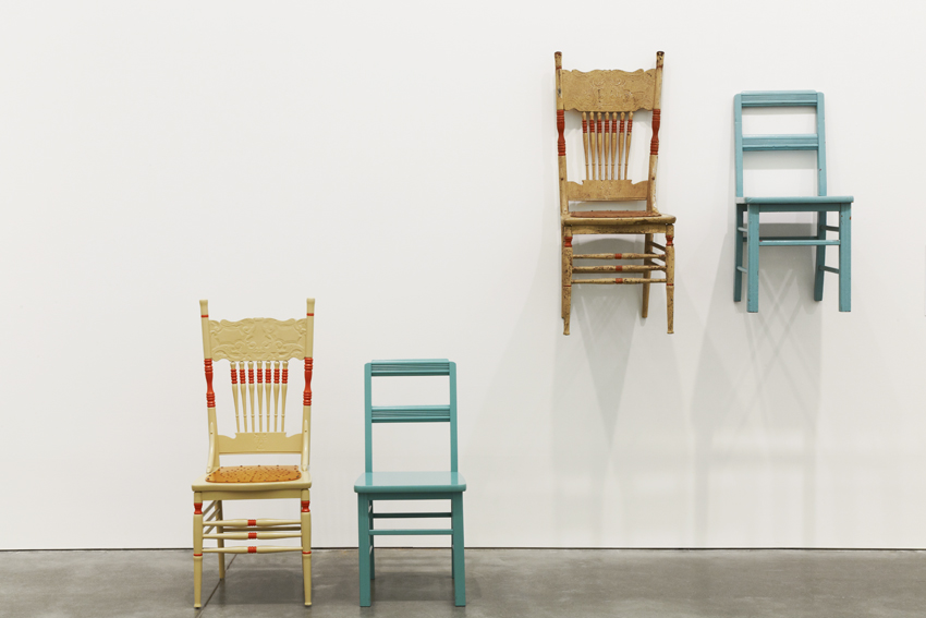 A sculptural installation of two identical pairs of chairs, one yellow and one blue chair. One pair is hung on the wall, one is on the floor.