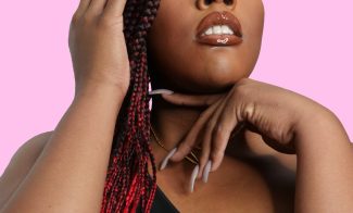 A glamour head shot of an African-American femme person with red and black braids, posing with their hands in front of a pink backdrop.