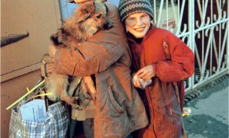 A color photograph shows a pale older woman holding a dog and a plastic bag of various materials, standing next to a pale school-age boy in a red jacket and black patterned knit hat who smiles directly at the viewer.