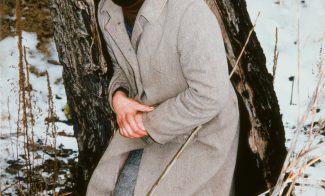 A color photograph shows a pale-skinned figure in a hat, hood, overcoat, and red boots hunched against a tree in a snowy forest, looking directly at the viewer.