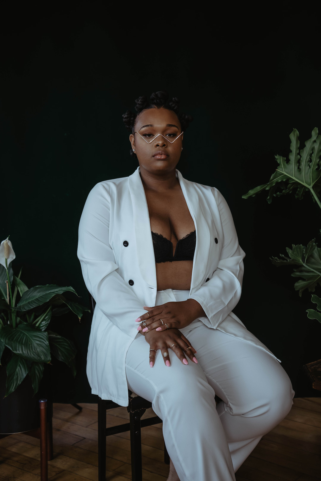 A seated portrait photo of a dark skinned woman with angular glasses in a white blazer and white pants in front of a black backdrop with plants.