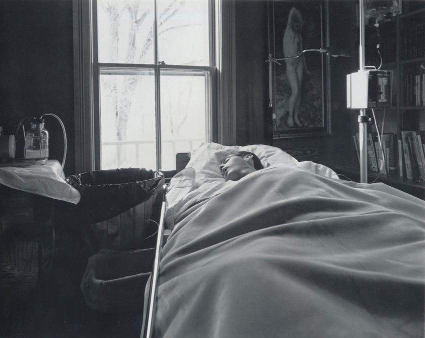 A black-and-white photograph of a man asleep under thick blankets in a home hospital bed, surrounded by an IV and other hospital equipment.