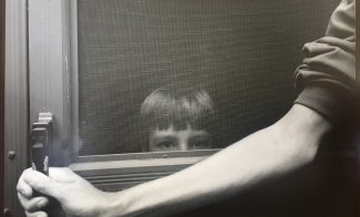 A black-and-white tinted photograph shows a pale, thin arm extended across a screen door, holding the handle, as a small child with bangs looks directly at the viewer through the screen.