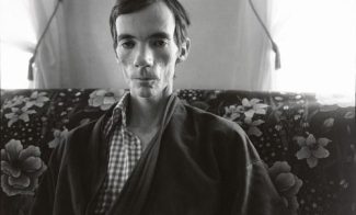 A black-and-white photograph of a very gaunt man sitting upright on a floral couch in a bathrobe.