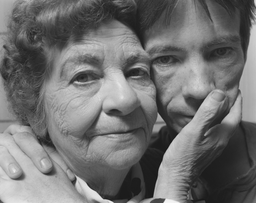 A black-and-white photograph of a light-skinned young man and his aging mother looking directly at the viewer, their faces touching.