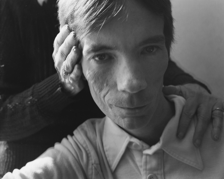 A black-and-white photograph of a pale-skinned man facing the viewer, as two hands touch the side of his face and shoulder.