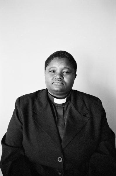 A black-and-white photograph of a person from the waist up wearing a blazer and clergy shirt against a blank wall