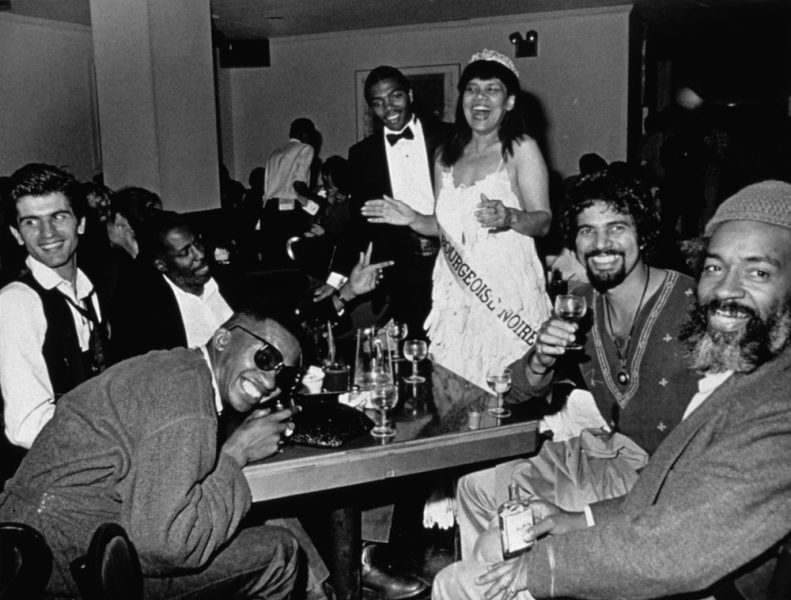 A black-and-white photograph of a Black woman in a beauty pageant gown, crown, and sash standing and laughing widely with her arms extended. In front of her are several men seated at a table full of drinks, smiling.