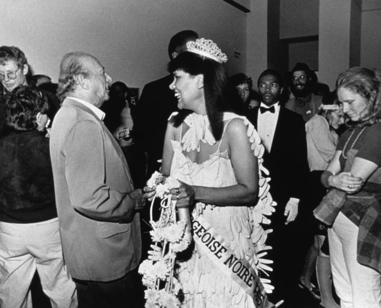 A black-and-white photograph of a Black woman in a beauty pageant gown, crown, gloves, bouquet, and sash in a crowded gallery, smiling at a someone unseen in the image.